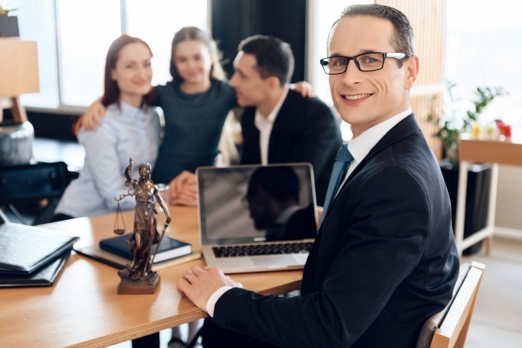 Smiling lawyer seated at desk with Lady Justice statue, laptop, and documents; team of professionals conversing in background